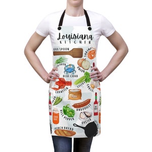 May include: White kitchen apron with a black strap and a colorful illustration of Louisiana kitchen ingredients, including shrimp, crawfish, okra, andouille sausage, and French bread. The text "Louisiana Kitchen" is printed in black at the top of the apron.