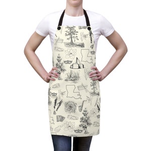 May include: A cream-colored apron with a black adjustable strap and black buttons. The apron features a black and white illustration of Louisiana landmarks, including the state outline, a cypress tree, a fleur-de-lis, a boat, a mask, and a musical instrument.