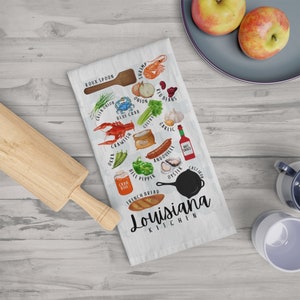 May include: A white kitchen towel with a colorful illustration of Louisiana cuisine ingredients, including shrimp, crawfish, okra, andouille sausage, and a cast iron skillet. The text "Louisiana Kitchen" is printed in black at the bottom of the towel.