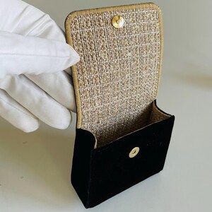 Op de afbeelding: Een kleine, open zwarte fluwelen tas met een goudkleurige sluiting. De binnenkant is gevoerd met een textiel van goud en beige. De tas wordt opengehouden en toont de binnenkant.