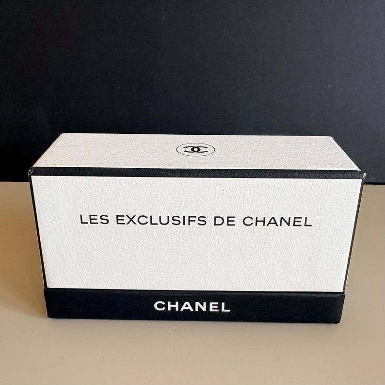 May include: A rectangular white box with black trim and the text "LES EXCLUSIFS DE CHANEL" and "CHANEL" printed on it. The top of the box features the Chanel logo. The box is on a light beige surface.
