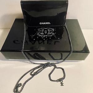 Puede incluir: Un bolso de cadena Chanel negro brillante con una correa de cadena plateada. El bolso est&aacute; sobre una caja negra con el logo de Chanel. El bolso tiene un cierre de solapa y el logo de Chanel es visible en la parte delantera.