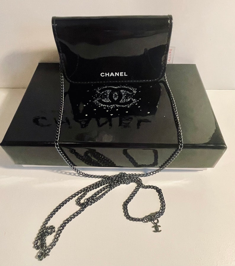 Puede incluir: Un bolso de cadena Chanel negro brillante con una correa de cadena plateada. El bolso est&aacute; sobre una caja negra con el logo de Chanel. El bolso tiene un cierre de solapa y el logo de Chanel es visible en la parte delantera.