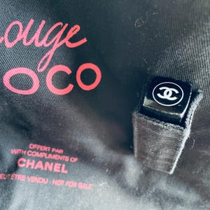 May include: Black lipstick with a white Chanel logo on the top. The lipstick is in a black fabric holder. The background is black with the words "Rouge Coco" in pink and the text "Offert par with compliments of Chanel".