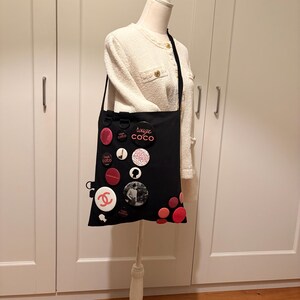 May include: Black tote bag adorned with various round, pink, and white decorative pins. The pins feature text and graphic designs. The bag is suspended from a mannequin wearing a white jacket.