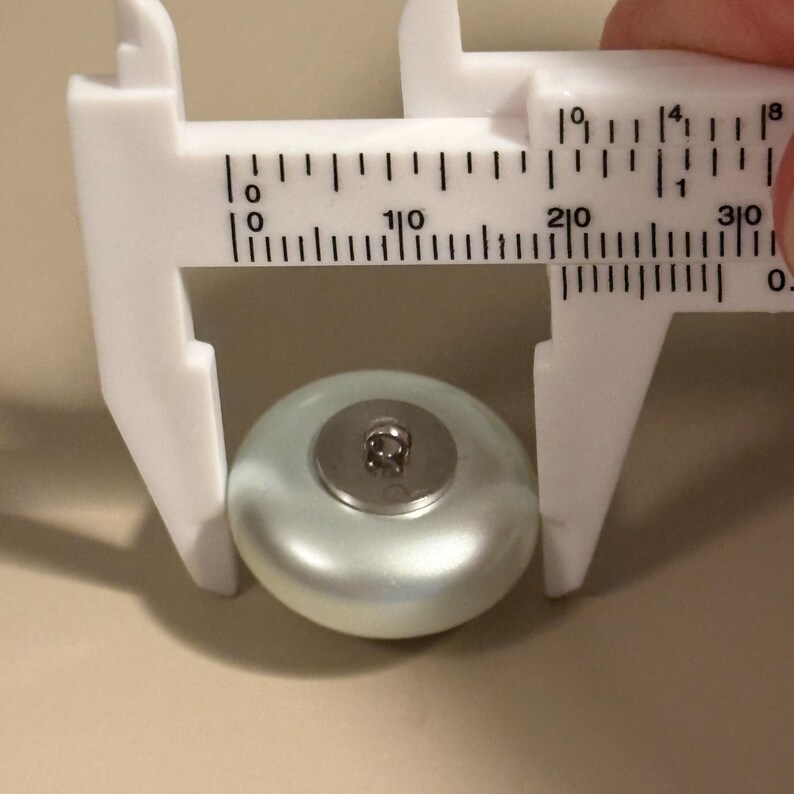 May include: A light green, pearlescent button with a metal shank is measured with a white caliper. The button measures approximately 0.75 inches in diameter. The button has a shiny, smooth surface.