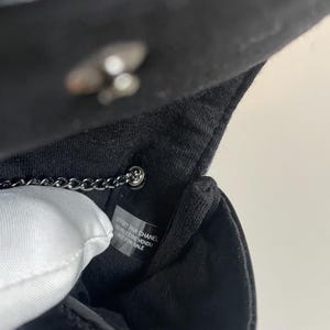 Puede incluir: Primer plano del interior de un bolso negro con una correa de cadena plateada. El interior del bolso presenta un forro blanco y una etiqueta con texto. El bolso est&aacute; hecho de charol brillante.