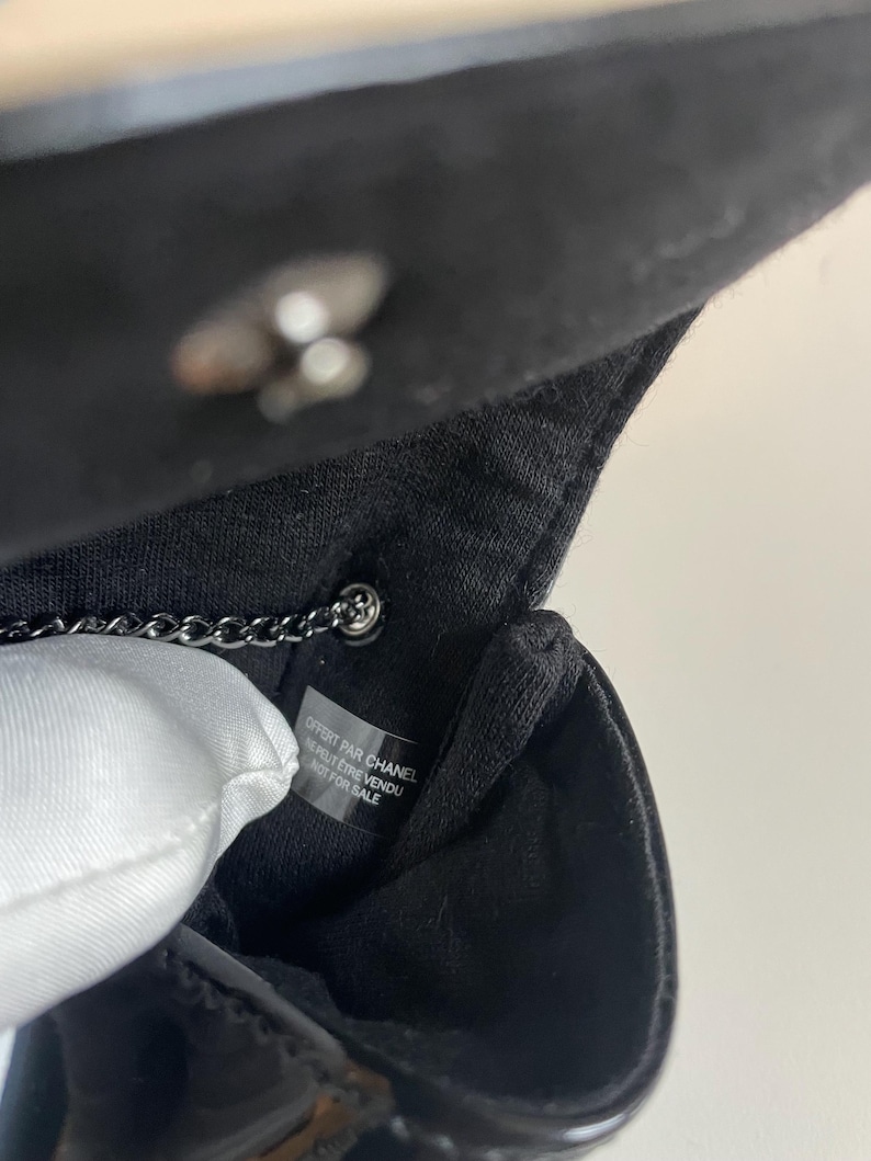 Puede incluir: Primer plano del interior de un bolso negro con una correa de cadena plateada. El interior del bolso presenta un forro blanco y una etiqueta con texto. El bolso est&aacute; hecho de charol brillante.