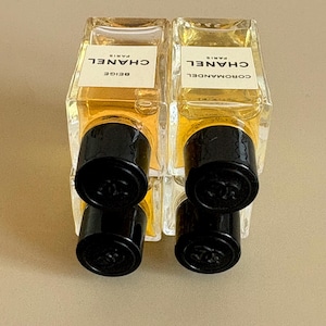May include: Two rectangular perfume bottles with black caps. The bottles are filled with a light amber liquid and have white labels with black text that reads "CHANEL PARIS". One bottle is labeled "BEIGE", the other "COROMANDEL".