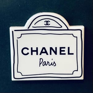 Könnte beinhalten: Ein weißes Schild mit schwarzem Rand und dem Text "CHANEL Paris" mit einem Doppel-C-Logo oben.
