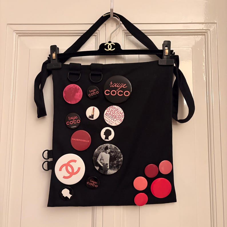 May include: Black tote bag with various round pins and buttons. The pins feature the words "rouge coco", "Mademoiselle", and a silhouette. The bag is hanging on a black hanger with a logo.