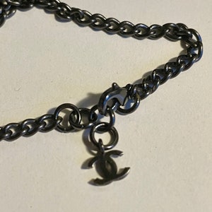 Puede incluir: Primer plano de un collar de cadena de color metal de ca&ntilde;&oacute;n. El collar presenta un dise&ntilde;o de cadena de bordillo y un peque&ntilde;o colgante con un logotipo de doble C. El cierre y el colgante son visibles sobre un fondo neutro.