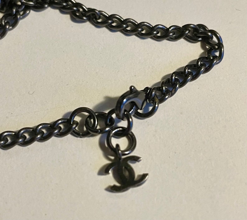 Puede incluir: Primer plano de un collar de cadena de color metal de ca&ntilde;&oacute;n. El collar presenta un dise&ntilde;o de cadena de bordillo y un peque&ntilde;o colgante con un logotipo de doble C. El cierre y el colgante son visibles sobre un fondo neutro.