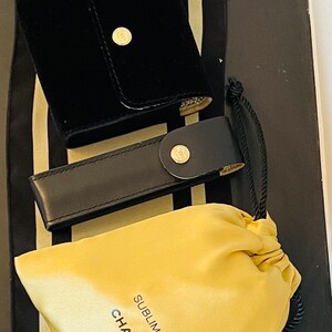 Op de afbeelding: Zwarte fluwelen etui met een gouden knoopsluiting, een zwarte leren etui met een gouden knoopsluiting en een gele trekkoordtas met de tekst "SUBLIMAGE CHANEL". De items staan op een zwart-wit gestreepte achtergrond.