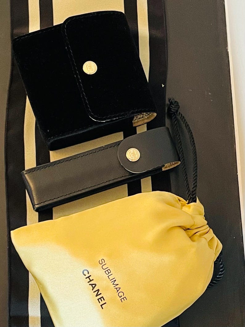 Op de afbeelding: Zwarte fluwelen etui met een gouden knoopsluiting, een zwarte leren etui met een gouden knoopsluiting en een gele trekkoordtas met de tekst "SUBLIMAGE CHANEL". De items staan op een zwart-wit gestreepte achtergrond.