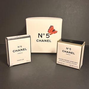 May include: Three Chanel N°5 products are displayed on a dark surface. The items include a white box with a red butterfly graphic and the text "N°5 CHANEL", and two smaller boxes with the same branding. One box is labeled "PARFUM", the other "POUDRE PRESSEE".