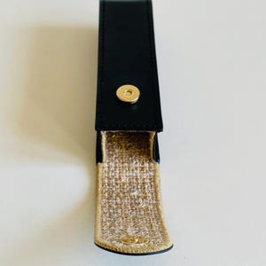 Op de afbeelding: Een zwart leren etui met een gouden drukknopsluiting. De binnenkant is gevoerd met een goud-beige geweven stof. De etui is ontworpen om een klein item in te bewaren, zoals een pen of lippenstift.