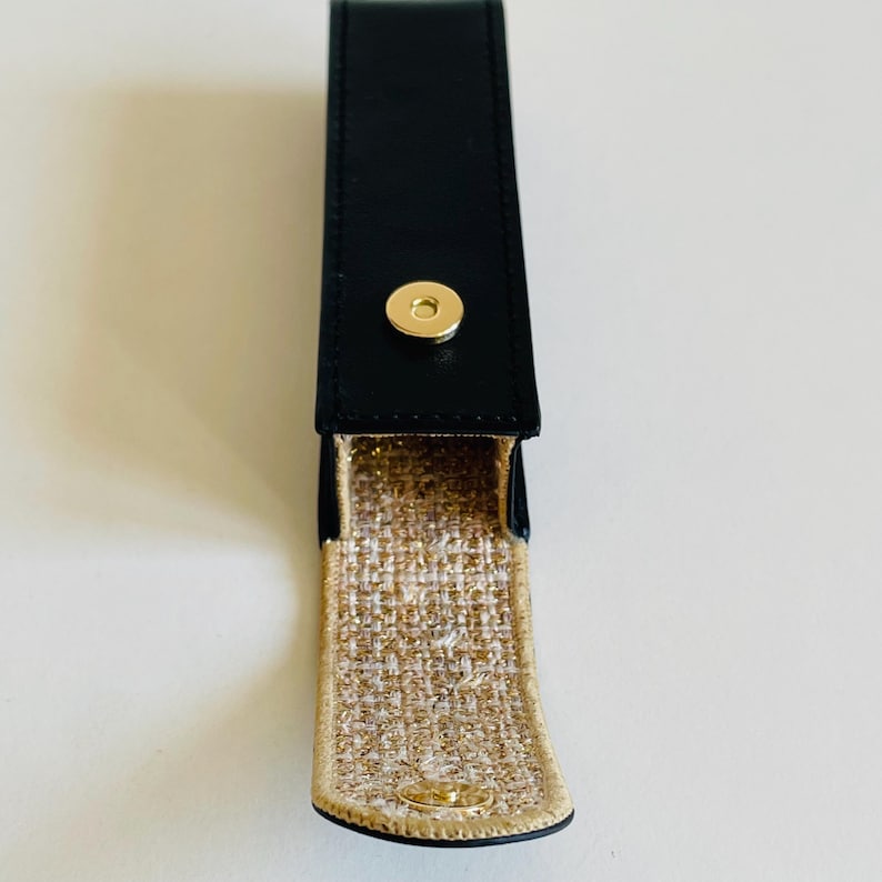 Op de afbeelding: Een zwart leren etui met een gouden drukknopsluiting. De binnenkant is gevoerd met een goud-beige geweven stof. De etui is ontworpen om een klein item in te bewaren, zoals een pen of lippenstift.