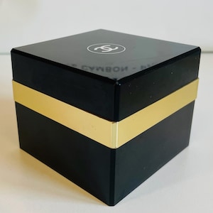 May include: A black square box with a gold band around the middle. The top of the box is glossy and features a white circular logo with the letters "S" and "C" intertwined. The box is likely for jewelry or a similar luxury item.
