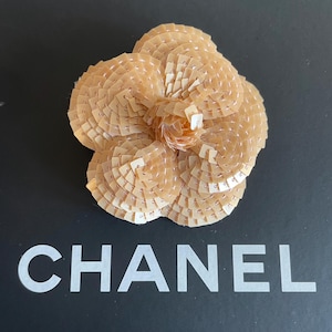 Puede incluir: Un broche grande de color beige en forma de flor hecho de lentejuelas. El broche está sobre un fondo negro con la palabra "CHANEL" impresa en blanco.