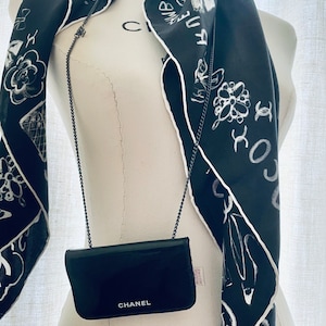 Puede incluir: Un bolso bandolera CHANEL negro con una correa de cadena se muestra en un maniqu&iacute;. Una bufanda de seda negra con ilustraciones y texto en blanco est&aacute; drapeada alrededor del cuello del maniqu&iacute;. La bufanda tiene un borde blanco.