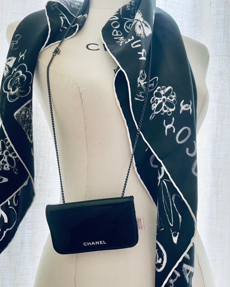 Puede incluir: Un bolso bandolera CHANEL negro con una correa de cadena se muestra en un maniqu&iacute;. Una bufanda de seda negra con ilustraciones y texto en blanco est&aacute; drapeada alrededor del cuello del maniqu&iacute;. La bufanda tiene un borde blanco.