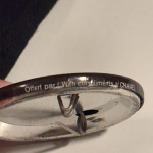 May include: A silver-colored pin with a dark brown border. The pin has the text "Offert par With compliments CHANEL" printed on the border. The pin has a metal clasp on the back.