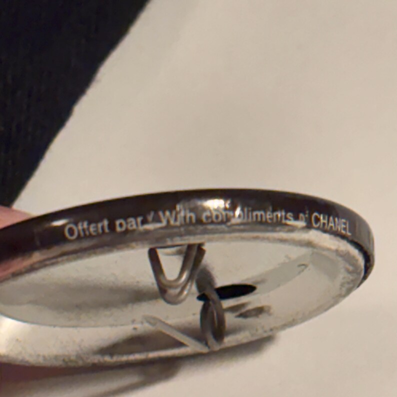 May include: A silver-colored pin with a dark brown border. The pin has the text "Offert par With compliments CHANEL" printed on the border. The pin has a metal clasp on the back.
