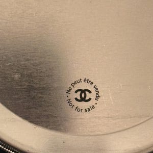 May include: Circular silver-toned object with the text "Ne peut &ecirc;tre vendu" and "Not for sale" surrounding a double-C logo. The object has a brushed metal finish and a dark border.