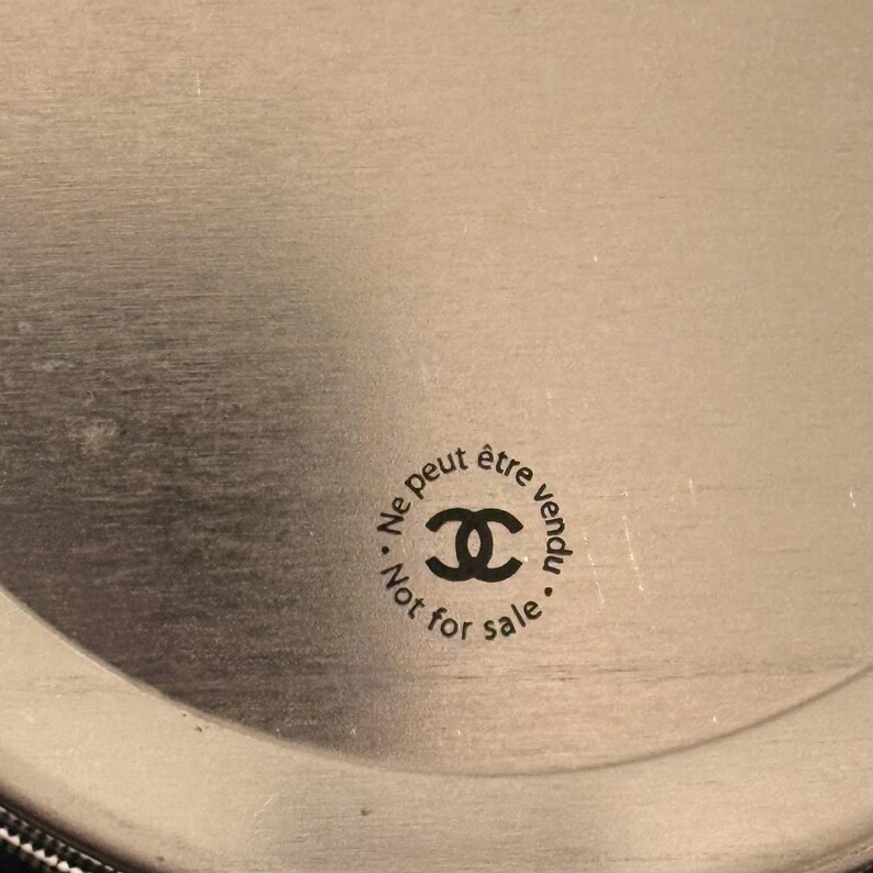 May include: Circular silver-toned object with the text "Ne peut &ecirc;tre vendu" and "Not for sale" surrounding a double-C logo. The object has a brushed metal finish and a dark border.