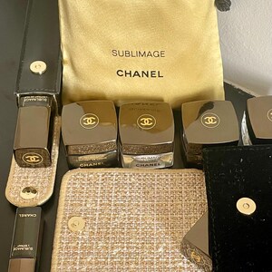 Op de afbeelding: Een assortiment Chanel Sublimage huidverzorgingsproducten. De afbeelding toont verschillende donkerbruine potten en tubes met gouden accenten en het Chanel-logo. Een gouden trekkoordtas met het Chanel-logo en tekst is ook aanwezig, samen met een kleine gouden en beige make-uptas.