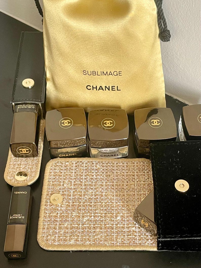 Op de afbeelding: Een assortiment Chanel Sublimage huidverzorgingsproducten. De afbeelding toont verschillende donkerbruine potten en tubes met gouden accenten en het Chanel-logo. Een gouden trekkoordtas met het Chanel-logo en tekst is ook aanwezig, samen met een kleine gouden en beige make-uptas.