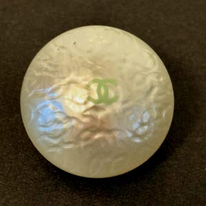 May include: A round, off-white button with a pearlescent finish. The button features a green logo in the center. The button is set against a dark background.
