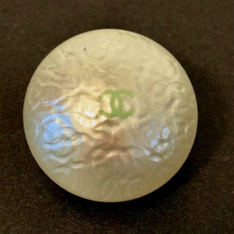 May include: A round, off-white button with a pearlescent finish. The button features a green logo in the center. The button is set against a dark background.