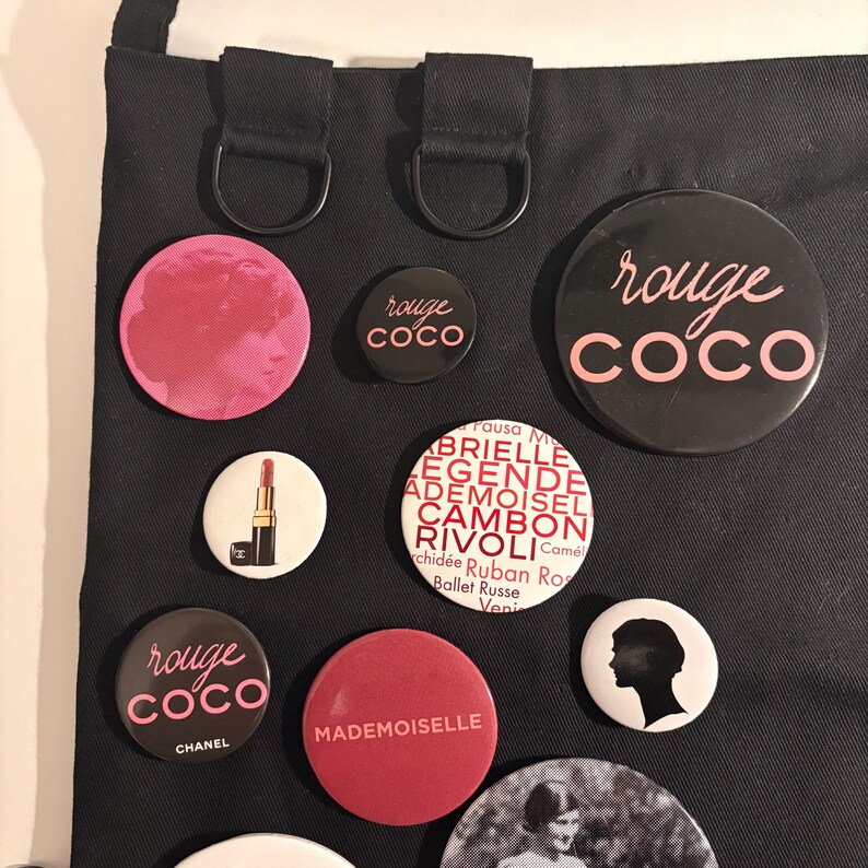 May include: A collection of round pins with various designs and text, including "rouge COCO" in pink and white, a lipstick, and the word "MADEMOISELLE". The pins are displayed on a black background.