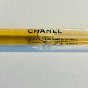 May include: A clear, slender perfume vial with a golden-yellow liquid inside. The vial has the word "CHANEL" printed in black, along with "ELIXIR SENSUEL" and other text. The vial is on a white surface.