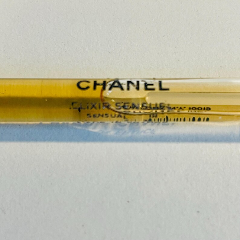 May include: A clear, slender perfume vial with a golden-yellow liquid inside. The vial has the word "CHANEL" printed in black, along with "ELIXIR SENSUEL" and other text. The vial is on a white surface.