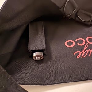 May include: A black handbag with a silver-colored clasp featuring a logo. The bag's interior is visible, with the clasp partially extended from a black strap. The word "CO" is visible in pink script.