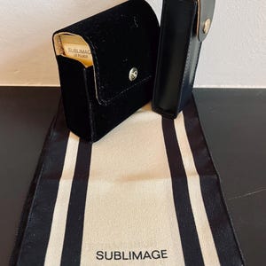 Op de afbeelding: Twee zwarte etuis, &eacute;&eacute;n van fluweel en &eacute;&eacute;n van leer, met gouden accenten, liggen op een cr&egrave;mekleurige stof met zwarte strepen. De fluwelen etui heeft een zilveren knoopsluiting. De stof toont de tekst "SUBLIMAGE PR&Eacute;CISION CHANEL".