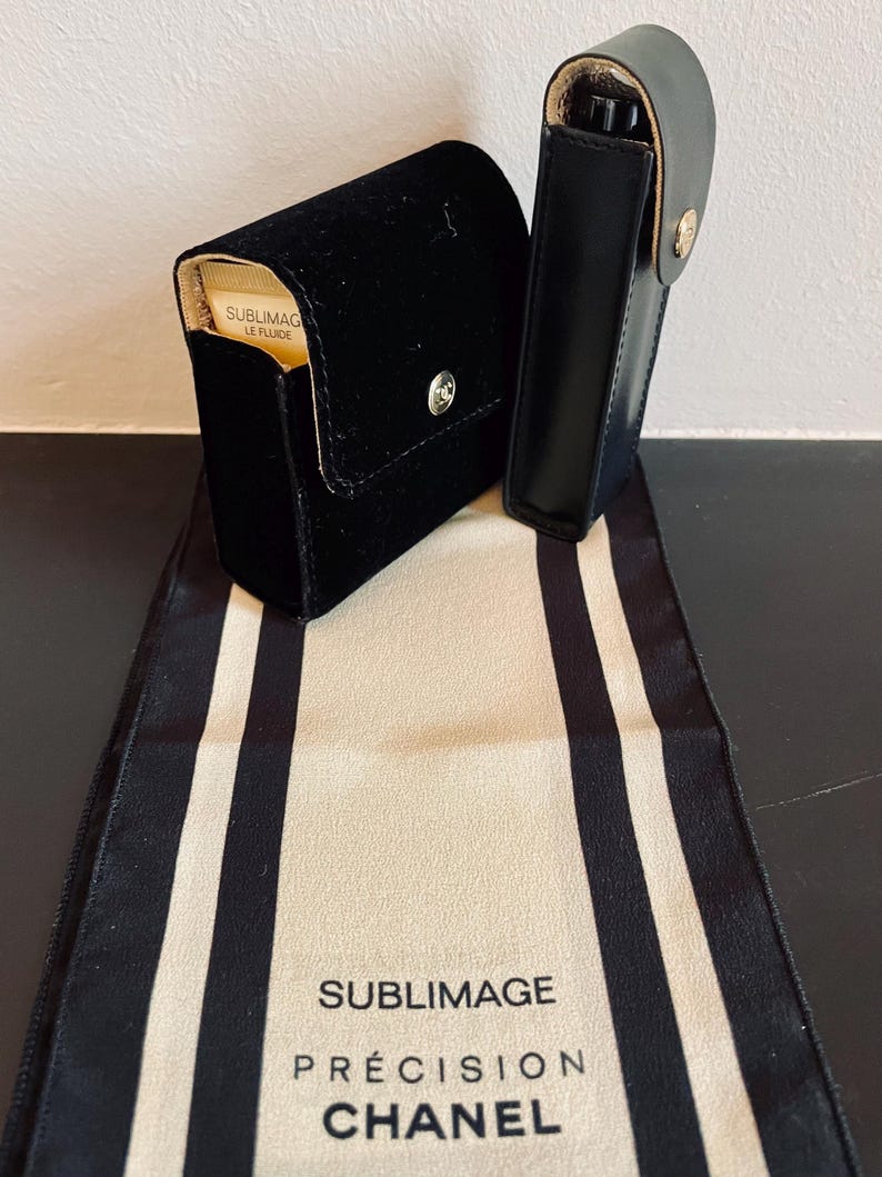 Op de afbeelding: Twee zwarte etuis, &eacute;&eacute;n van fluweel en &eacute;&eacute;n van leer, met gouden accenten, liggen op een cr&egrave;mekleurige stof met zwarte strepen. De fluwelen etui heeft een zilveren knoopsluiting. De stof toont de tekst "SUBLIMAGE PR&Eacute;CISION CHANEL".