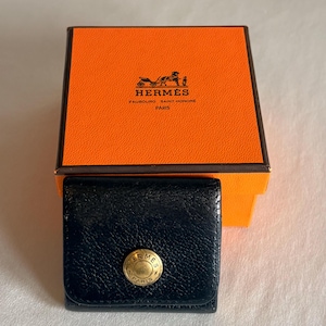 Puede incluir: Un monedero de cuero negro con un botón de presión circular dorado que presenta el texto HERMES PARIS. El monedero está colocado frente a una caja cuadrada naranja con borde marrón, que muestra el logotipo HERMES FAUBOURG SAINT-HONORE PARIS.
