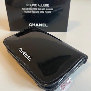 Puede incluir: Un mini bolso CHANEL negro brillante con el nombre de la marca en letras blancas. El bolso es rectangular con esquinas redondeadas. Una caja negra con las palabras "ROUGE ALLURE" y "CHANEL" est&aacute; en el fondo. Una peque&ntilde;a etiqueta de pl&aacute;stico transparente est&aacute; adherida al bolso.