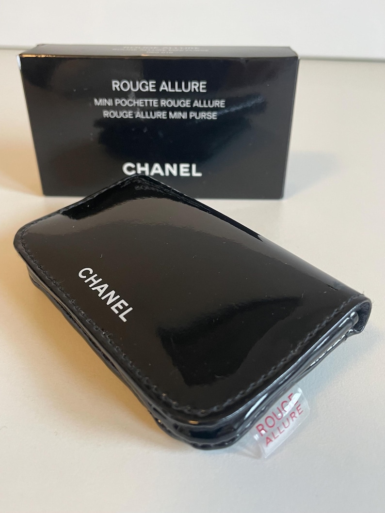 Puede incluir: Un mini bolso CHANEL negro brillante con el nombre de la marca en letras blancas. El bolso es rectangular con esquinas redondeadas. Una caja negra con las palabras "ROUGE ALLURE" y "CHANEL" est&aacute; en el fondo. Una peque&ntilde;a etiqueta de pl&aacute;stico transparente est&aacute; adherida al bolso.