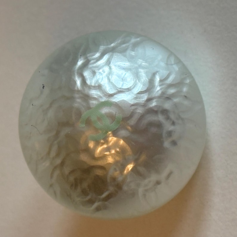 May include: A round, pearlescent button with a light green logo. The button has a textured surface and a subtle, swirling pattern. The logo is a stylized double C design.