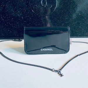Puede incluir: Una cartera de charol negro con una cadena plateada. La palabra "CHANEL" est&aacute; impresa en blanco en la parte delantera. Una bolsa negra m&aacute;s grande est&aacute; en el fondo.