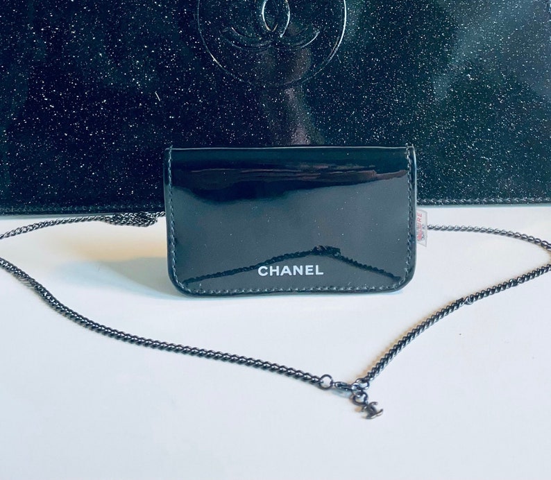 Puede incluir: Una cartera de charol negro con una cadena plateada. La palabra "CHANEL" est&aacute; impresa en blanco en la parte delantera. Una bolsa negra m&aacute;s grande est&aacute; en el fondo.