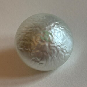 May include: A round, pearlescent button with a swirling, textured surface. A light green logo is visible on the surface. The button is likely for clothing or accessories.