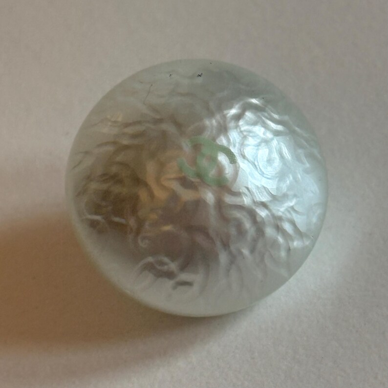 May include: A round, pearlescent button with a swirling, textured surface. A light green logo is visible on the surface. The button is likely for clothing or accessories.