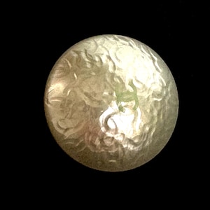 May include: A close-up of a round, metallic object with a textured surface. The object has a light, silvery color with subtle variations and a slightly irregular shape. The background is solid black.