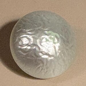 May include: A large, oval-shaped button with a pearlescent, iridescent finish. The button has a slightly textured surface and a subtle, swirling pattern. The color is primarily a soft, silvery-gray with hints of green and white.